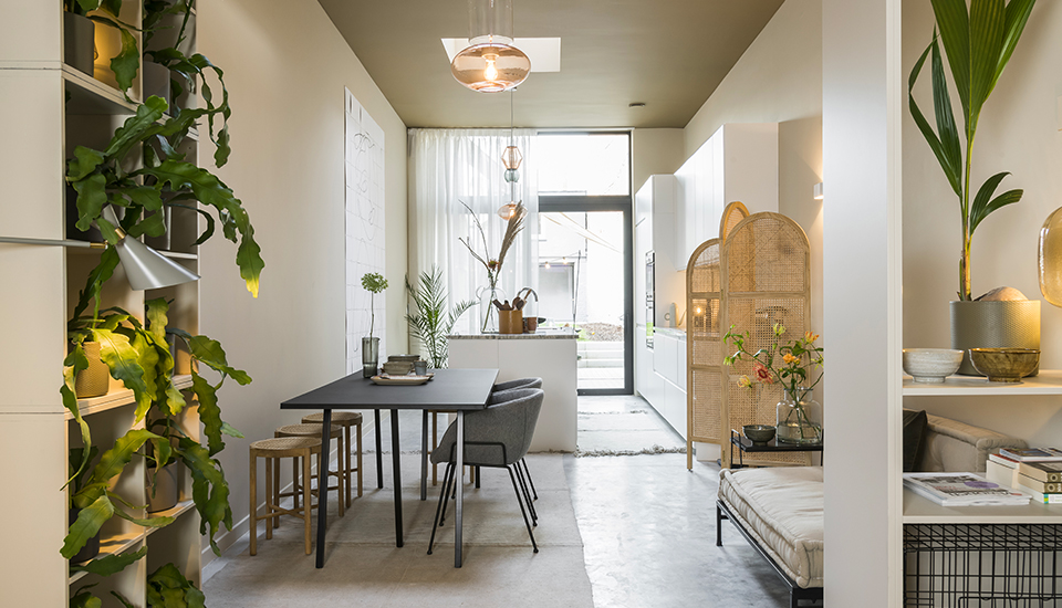 Salle à manger moderne avec table noire, chaises grises, tabourets en bois, plantes vertes et lumière naturelle entrant par une grande fenêtre.