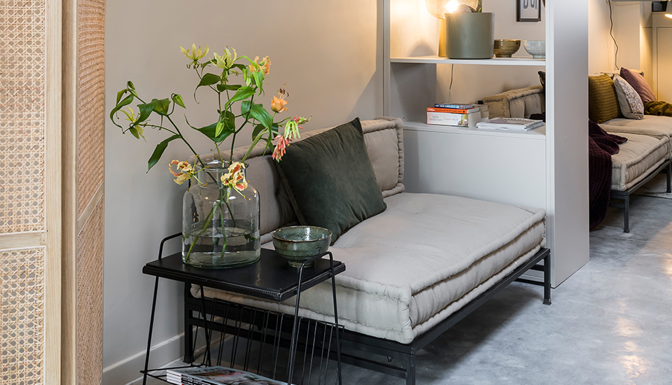 Un canapé beige avec coussin vert est placé à côté d'une table noire avec un vase en verre contenant des fleurs.