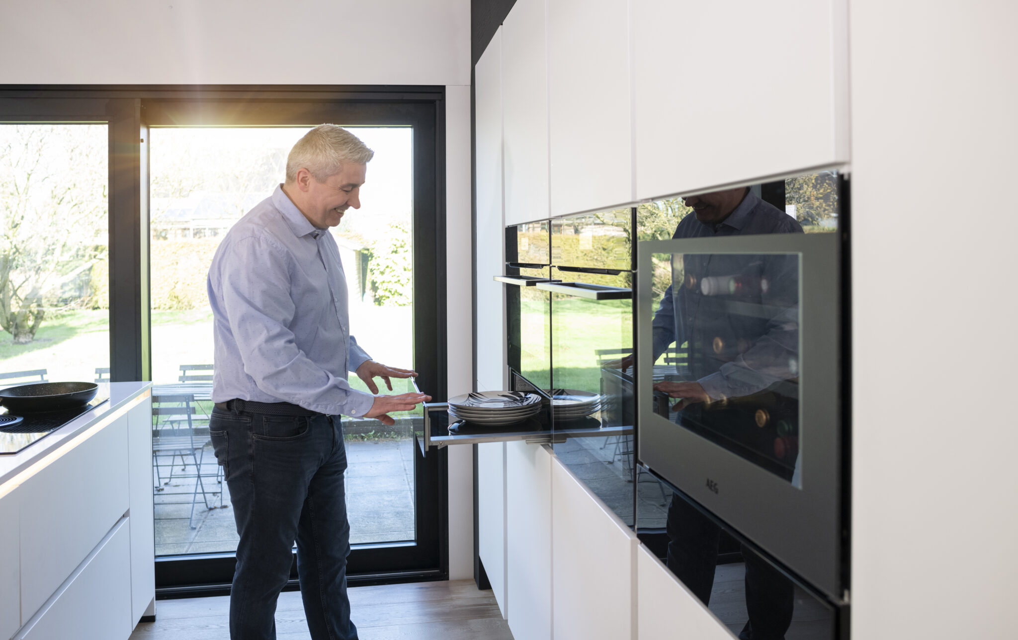Un homme en chemise bleue utilise un tiroir de cuisine moderne avec des assiettes, près d'une grande baie vitrée.