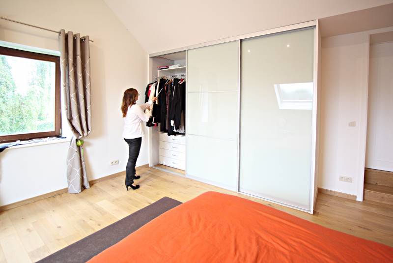 Une femme organise des vêtements dans un placard à portes coulissantes blanches, dans une chambre avec un lit à couverture orange.