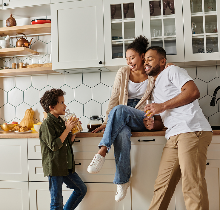 Une famille souriante se tient dans une cuisine moderne avec des placards blancs et un dosseret hexagonal, tenant des boissons.