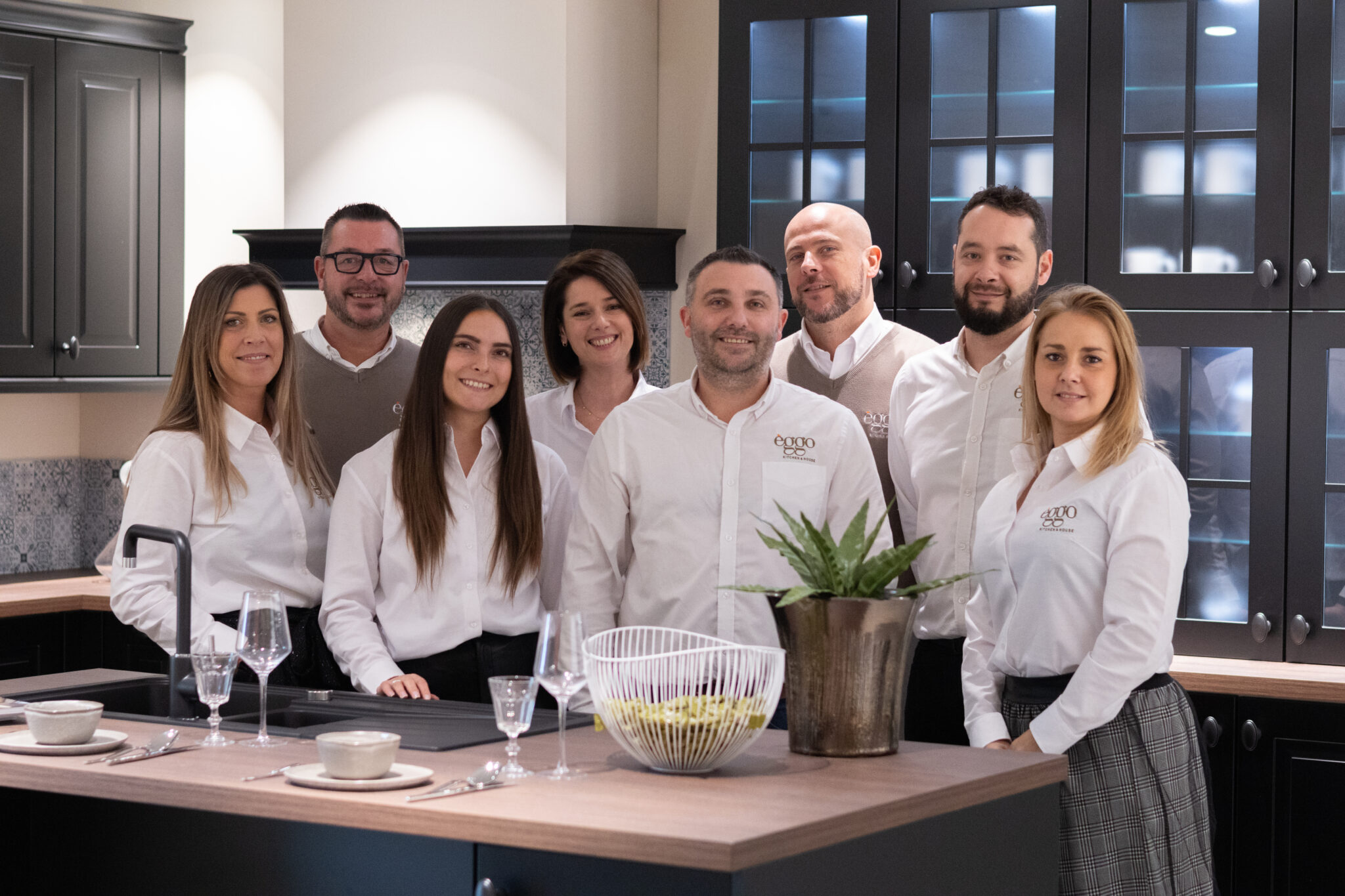 Un groupe de huit personnes en chemises blanches se tient dans une cuisine moderne avec des armoires noires et un plan de travail en bois.