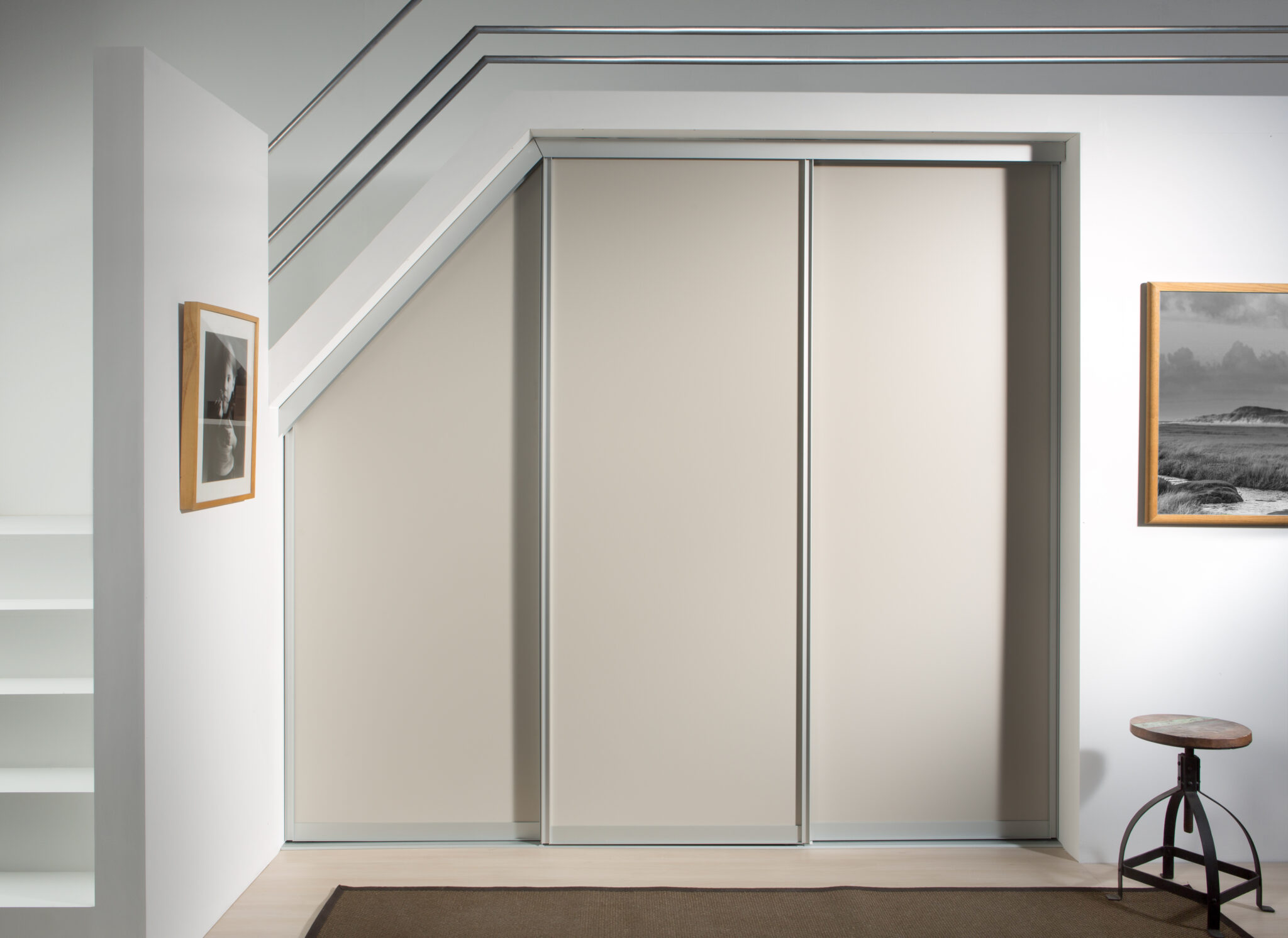 Porte coulissante beige sous un escalier, avec un tabouret en métal et bois, et deux cadres photo accrochés au mur blanc.