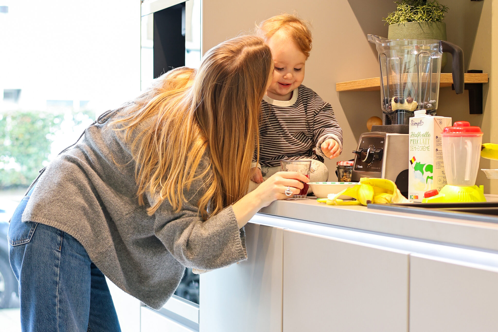 Une femme et un enfant interagissent dans une cuisine avec un mixeur, un verre de jus, des bananes, et une boîte de lait.