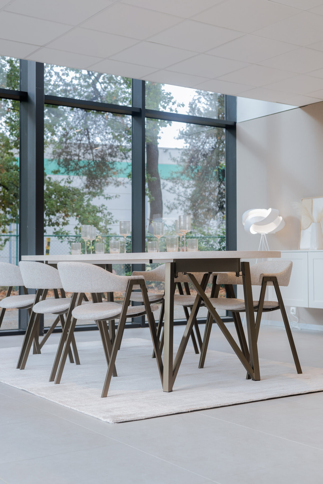 Une table en bois clair entourée de six chaises beige se trouve sur un tapis, devant une grande baie vitrée.