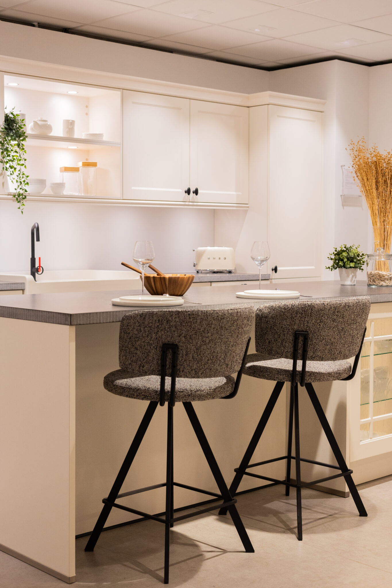 Cuisine moderne avec îlot central gris, deux chaises hautes grises, et étagères blanches décorées de plantes et vaisselle blanche.