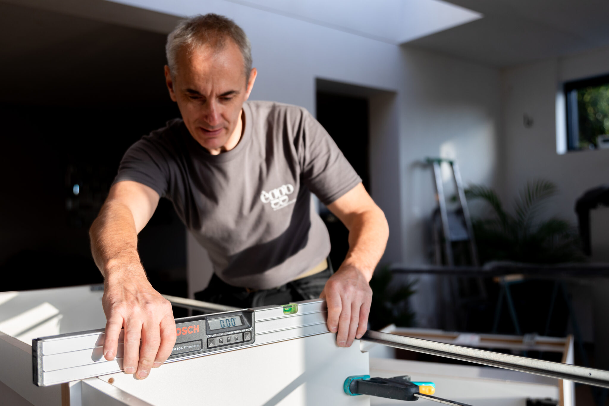 Homme mesurant un meuble blanc avec un niveau à bulle Bosch dans une pièce lumineuse, échelle et plantes en arrière-plan.
