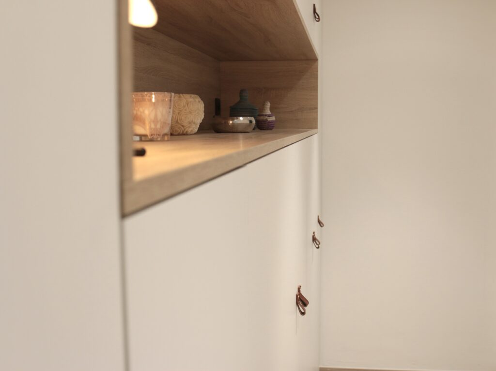 Étagère en bois clair avec des objets décoratifs, dont une bougie et un pot, au-dessus de placards blancs avec poignées en cuir marron.