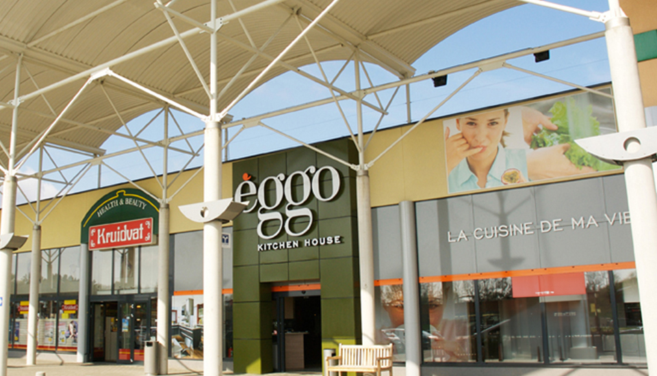Façade de magasins avec enseignes Kruidvat et èggo sous une structure métallique blanche, ciel bleu en arrière-plan.