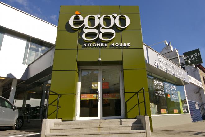 Façade d'un magasin èggo avec une entrée centrale vitrée et un panneau vert portant le logo blanc, entourée d'autres bâtiments.