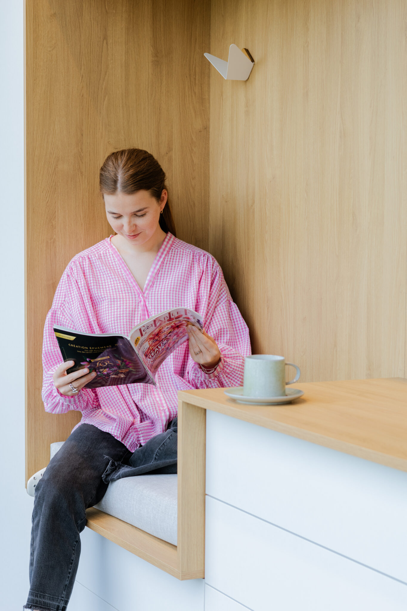 Une personne en chemise rose lit un magazine assise sur un banc en bois clair, avec une tasse verte posée à côté.