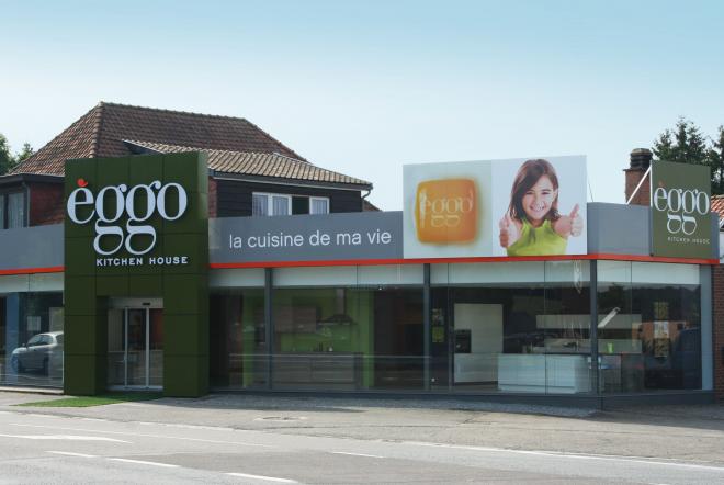 Façade d'un magasin èggo avec enseigne verte et affiches publicitaires, fenêtres vitrées, et maison en briques à l'arrière-plan.