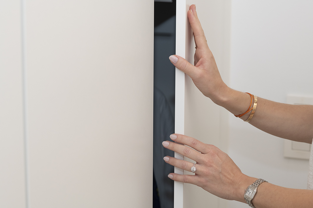 Des mains ouvrent une porte de placard blanche, montrant un intérieur sombre, avec des bracelets et une bague visibles.