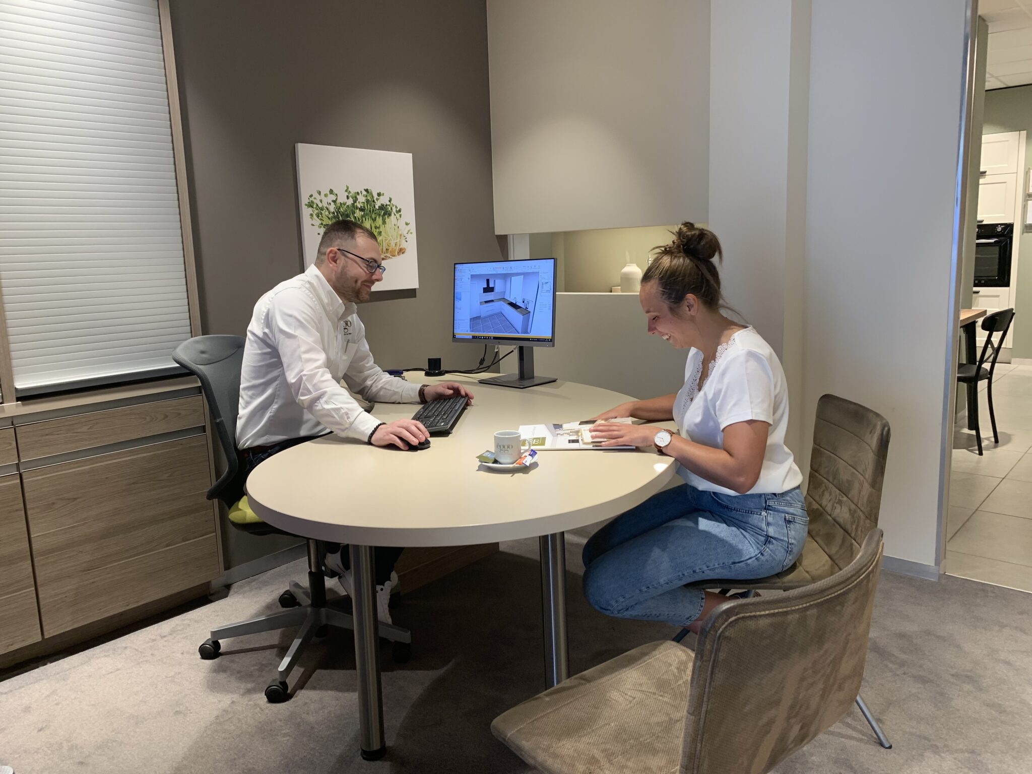 Deux personnes discutent à une table ronde dans un bureau, avec un ordinateur affichant un plan de cuisine.