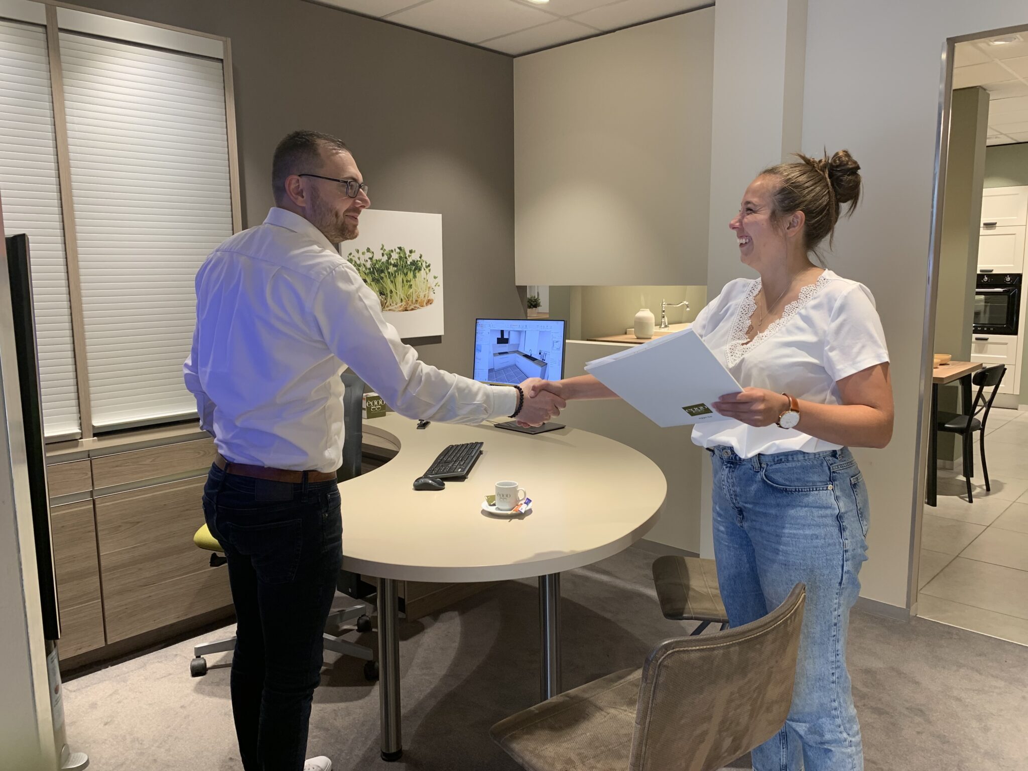 Deux personnes se serrent la main dans un bureau moderne avec un ordinateur, des dossiers et une tasse de café sur la table.