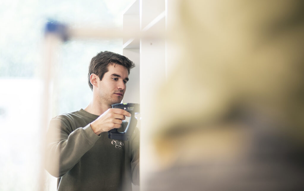Un homme en pull vert utilise une perceuse électrique pour monter un meuble blanc.