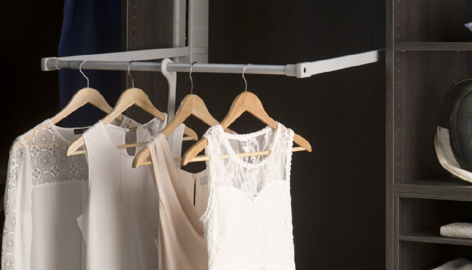 Des vêtements clairs sur cintres en bois sont suspendus à une tringle grise dans une penderie sombre, avec des étagères visibles à droite.
