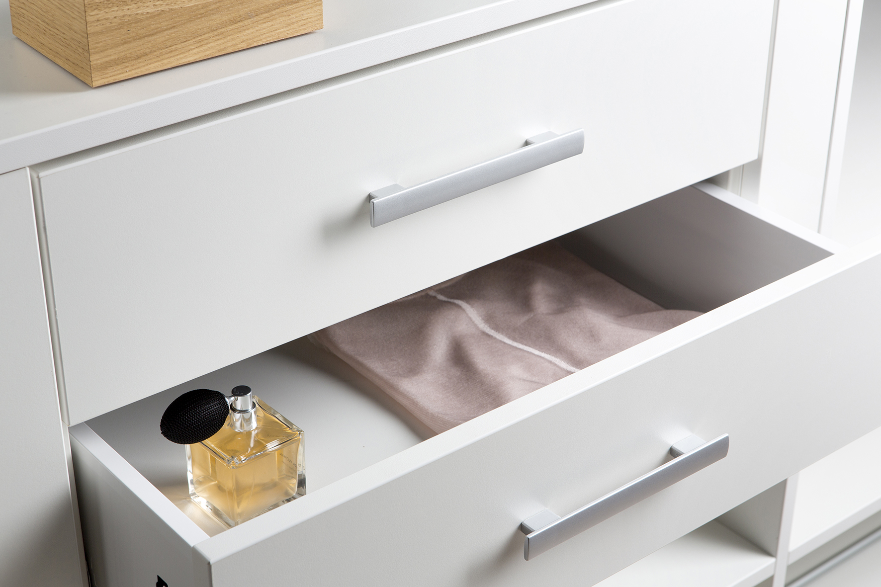 Un tiroir blanc ouvert contenant un tissu rose et un flacon de parfum carré avec un vaporisateur noir sur le côté.