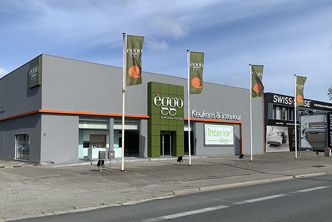 Façade grise du magasin èggo avec enseigne verte et orange, entourée de drapeaux, sur un trottoir pavé sous un ciel bleu.
