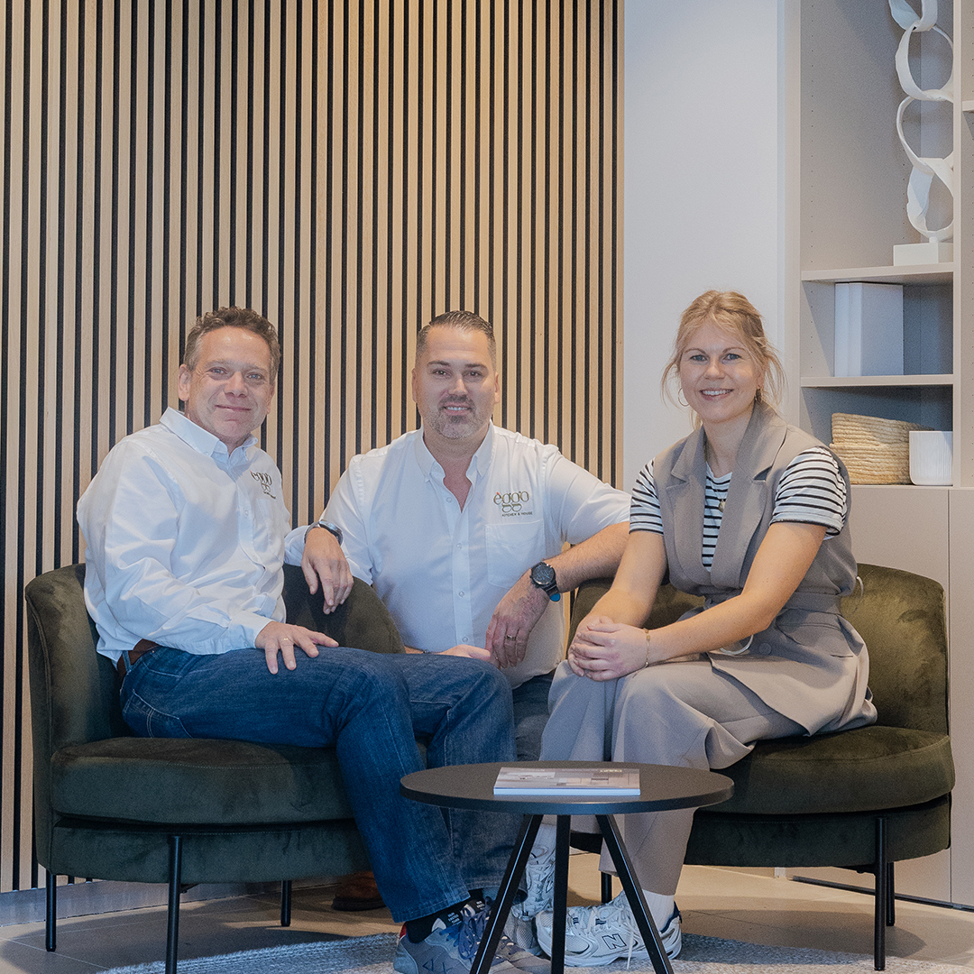 Trois personnes assises sur des fauteuils verts autour d'une table basse noire, devant un mur à rayures verticales.