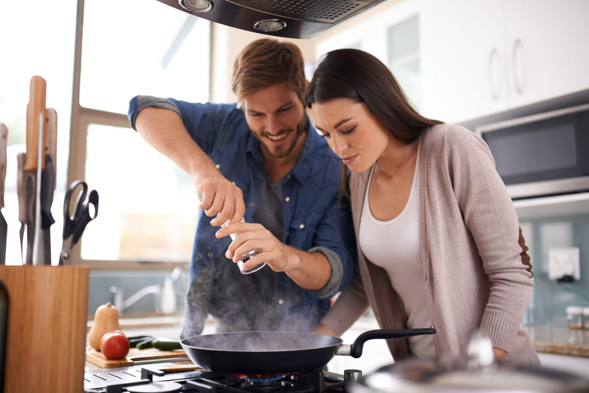 Un homme et une femme cuisinent ensemble, utilisant une poêle sur une cuisinière dans une cuisine moderne lumineuse.