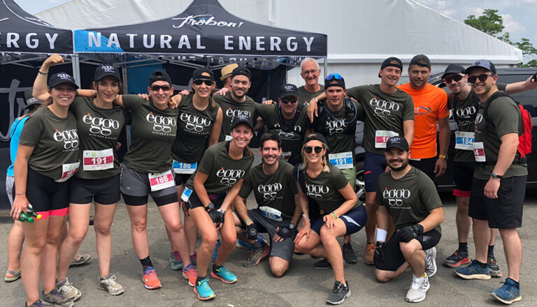Un groupe de personnes en t-shirts kaki avec le logo èggo sourit devant une tente marquée "Natural Energy".