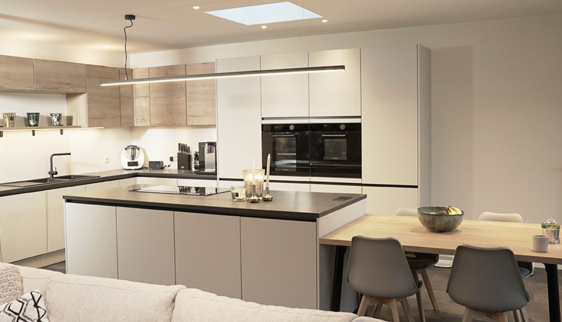 Cuisine moderne avec îlot central gris, meubles blancs et bois clair, équipée de fours encastrés, plan de travail noir et table à manger adjacente.