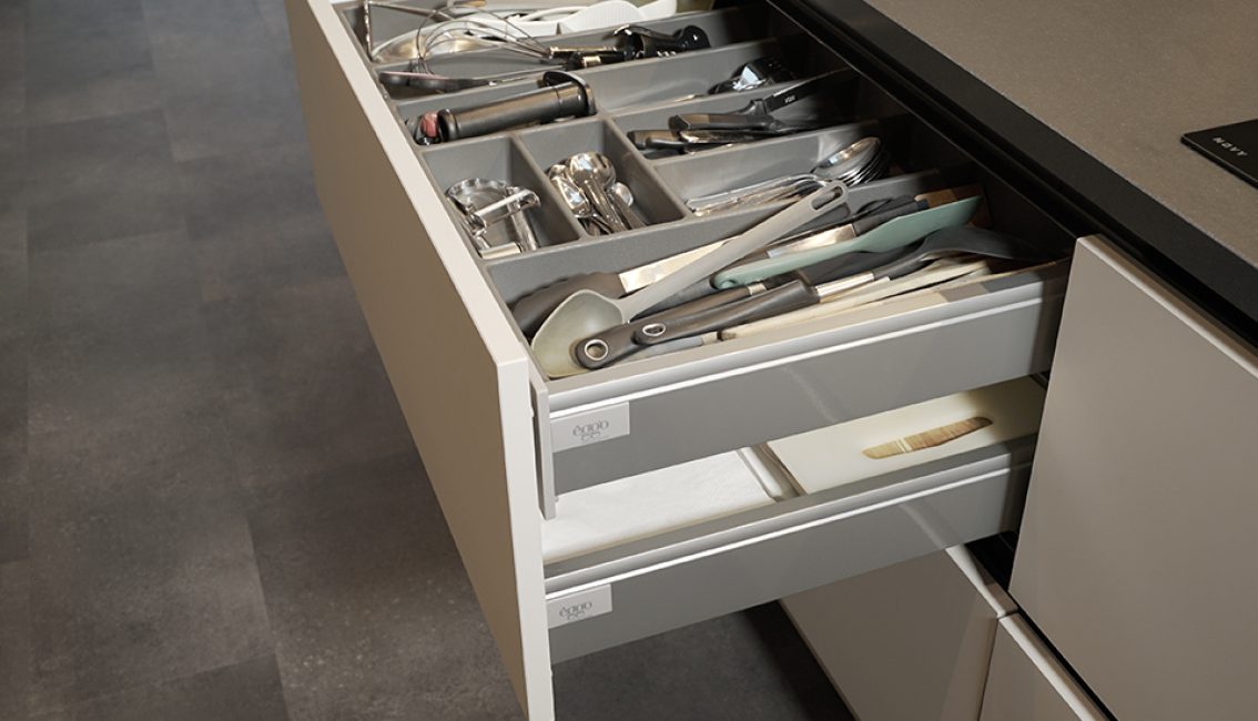 Un tiroir de cuisine ouvert contenant des ustensiles de cuisine en métal et plastique, avec un plan de travail gris visible au-dessus.