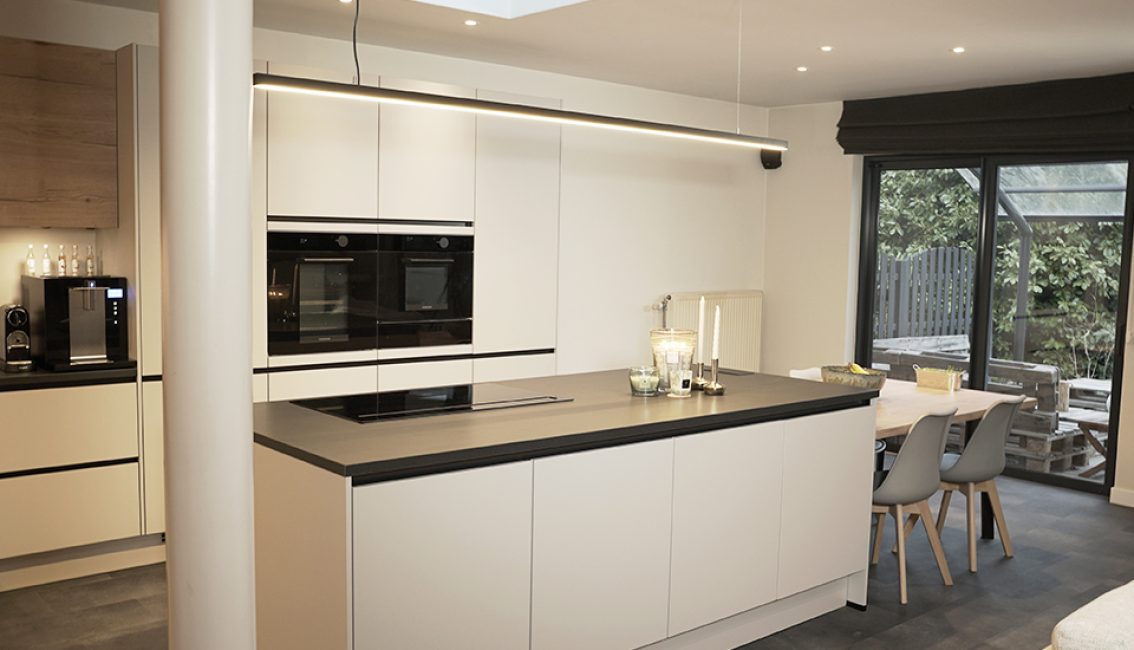 Cuisine moderne avec îlot central blanc, plan de travail noir, électroménager intégré, et table à manger avec chaises grises près d'une baie vitrée.