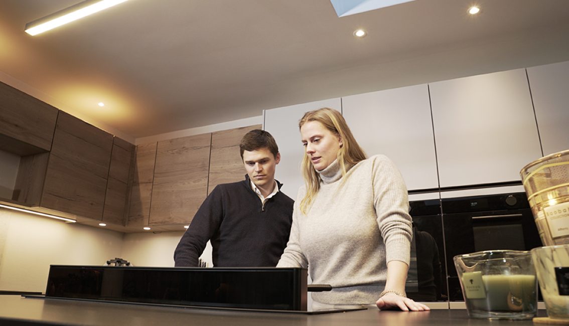 Deux personnes se tiennent dans une cuisine moderne avec des armoires en bois clair et un four encastré noir.