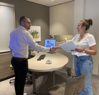 Deux personnes se serrent la main dans un bureau moderne avec un ordinateur, des dossiers et une tasse de café sur la table.